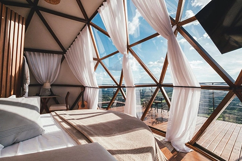 Quarto acolhedor com cortinas abertas, revelando a entrada suave da luz natural e a vista panorâmica da cabana