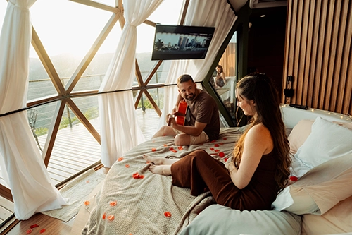 Momento de cumplicidade e música: casal relaxando na cama redonda enquanto o noivo toca violão