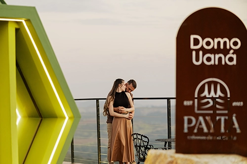 Casal abraçado e sorridente posando em frente à majestosa arquitetura exterior do Domo Uaná